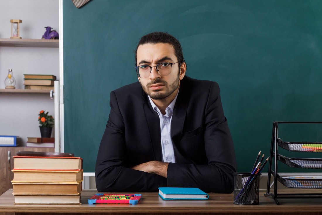 A tutor sitting at a desk in front of a chalkboard with teaching materials