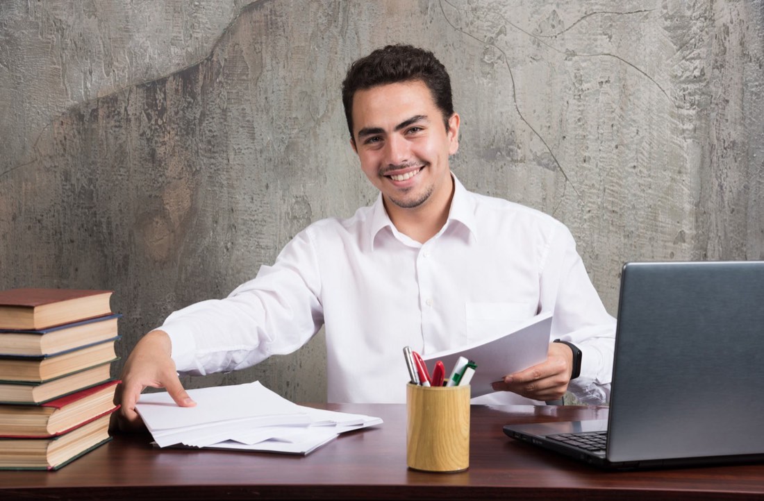 A tutor smiling at a desk with a laptop and books